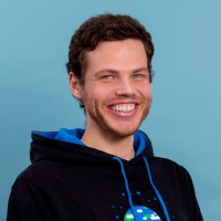 Portrait photo of a smiling man with dark curly hair wearing a hoodie against a blue background.