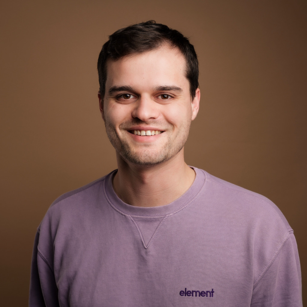 Portrait photo of a smiling man with short dark hair wearing a purple sweatshirt against a brown background.