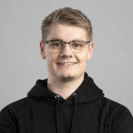 Portrait photo of a smiling man with short blond hair and glasses wearing a black hoodie against a gray background.
