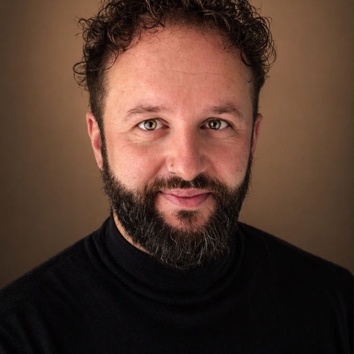Portrait photo of a smiling man with curly dark hair and a beard wearing a black turtleneck against a brown background.